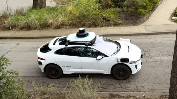 A white Waymo autonomous vehicle with a sensor on top drives along a residential street surrounded by greenery.