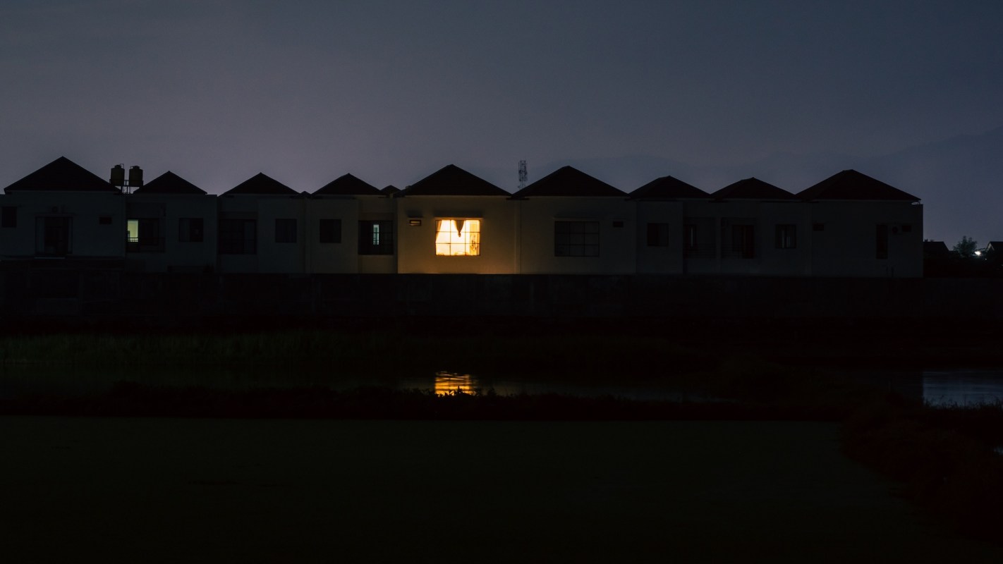A dark scene featuring a row of buildings with one illuminated window reflecting on the nearby water.