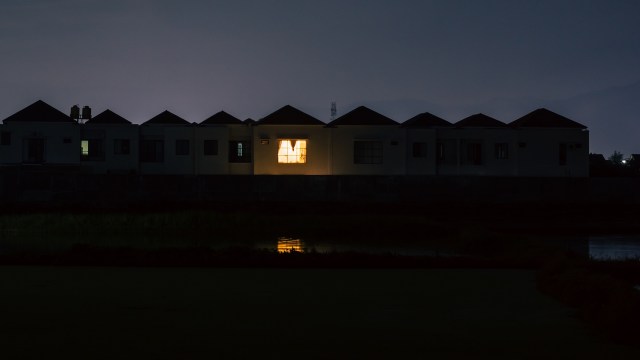 A dark scene featuring a row of buildings with one illuminated window reflecting on the nearby water.