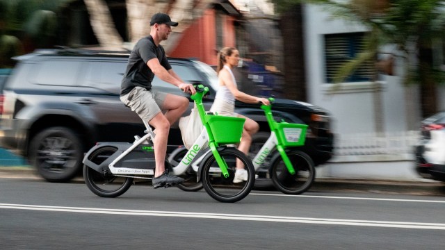 Two cyclists ride Lime electric bikes alongside a busy street, captured in motion.