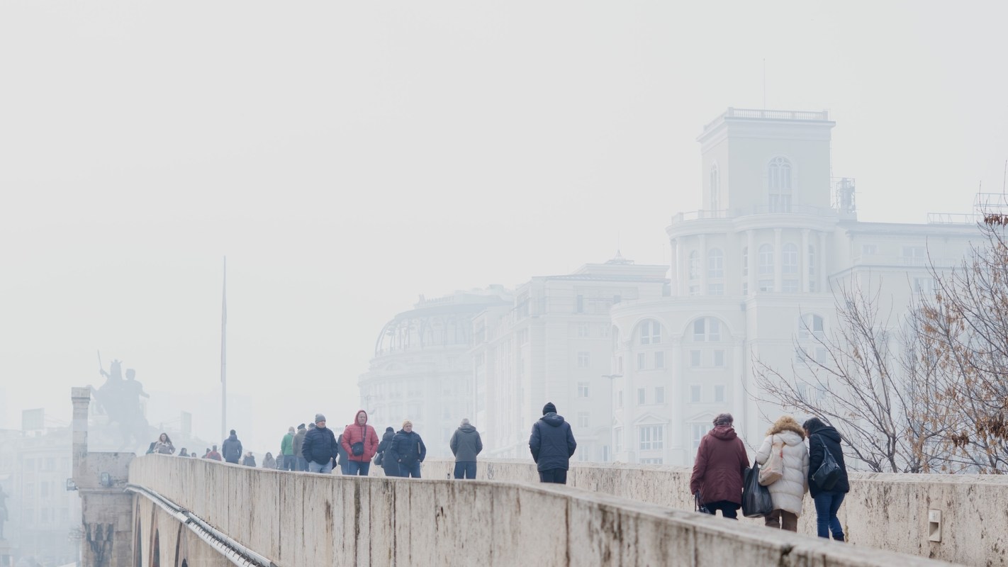 People walking across a bridge in a cityscape filled with smog with buildings barely visible in the background.