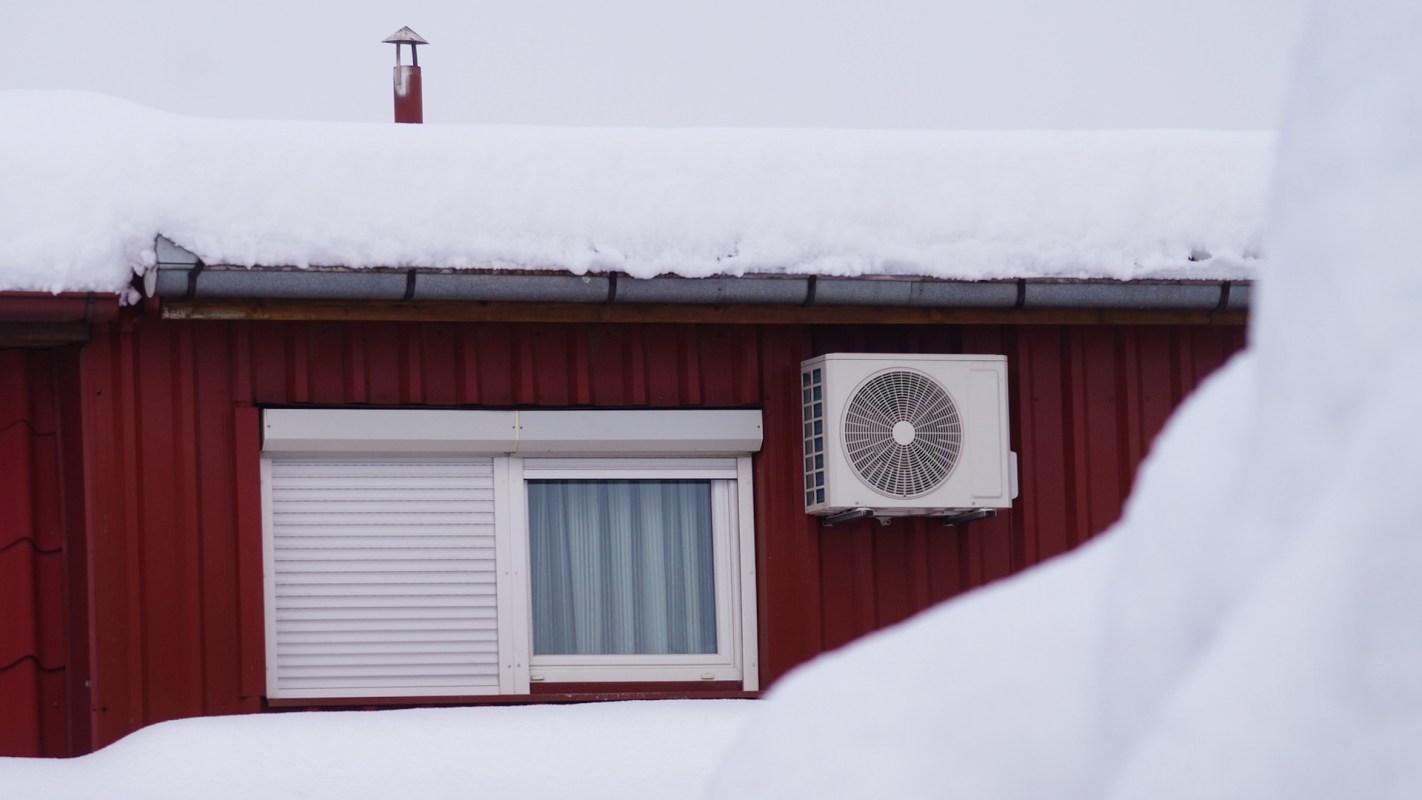 A red building with a snow-covered roof and an air conditioning unit near a partially obscured window.