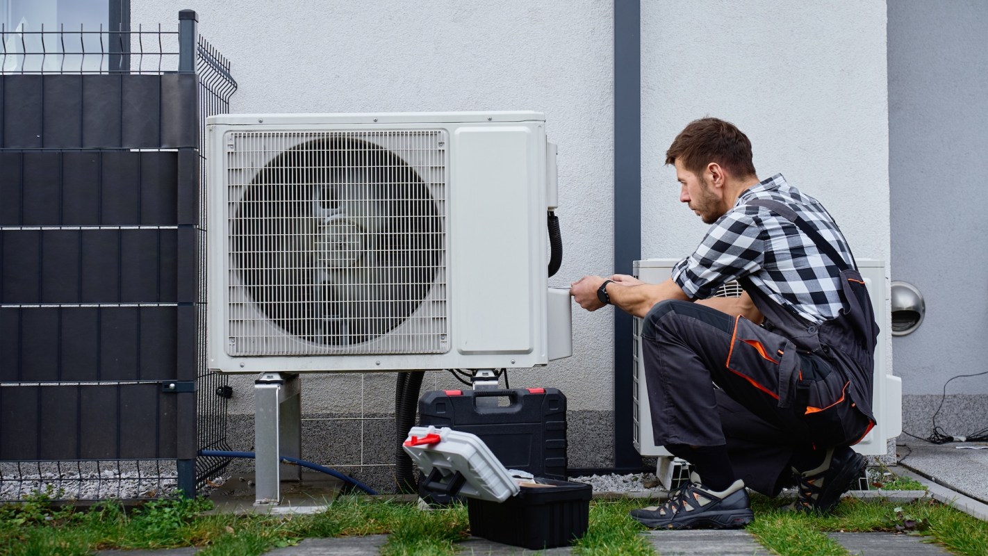 A technician crouches by a heat pump unit outside a home, checking tools and equipment on the ground.