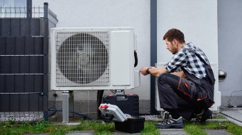 A technician crouches by a heat pump unit outside a home, checking tools and equipment on the ground.