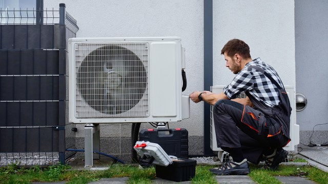 A technician crouches by a heat pump unit outside a home, checking tools and equipment on the ground.