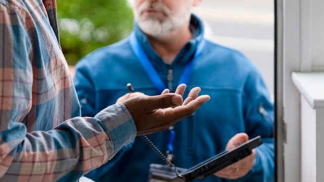 A person in a plaid shirt converses with a man holding a tablet at a doorstep.
