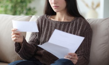 A woman in a brown sweater sits on a couch, holding and reading letters with a focused expression.
