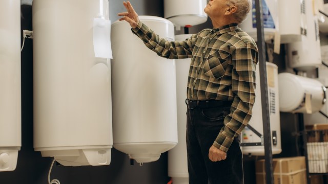 An elderly man in a plaid shirt examines white water heaters in a home appliance store.