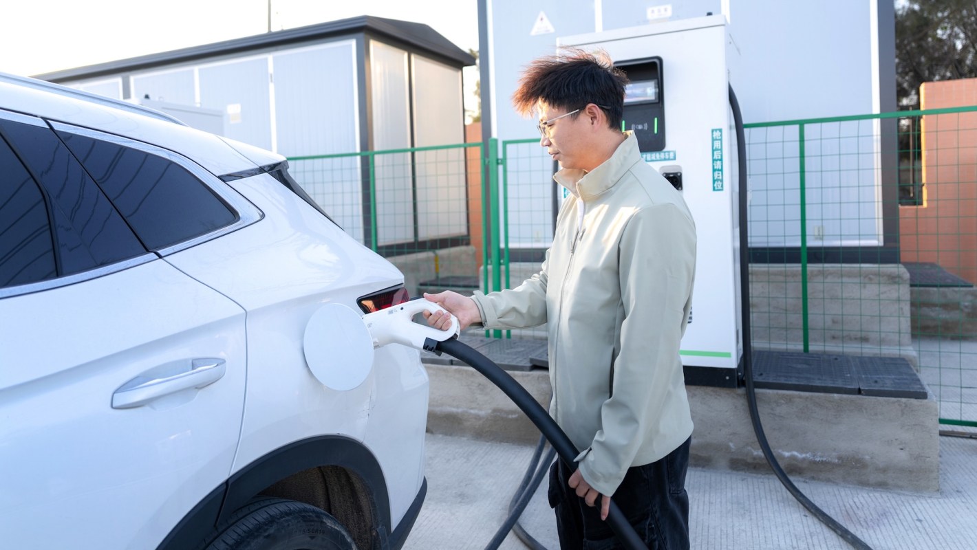 A person charges an electric vehicle at a charging station in an outdoor setting.