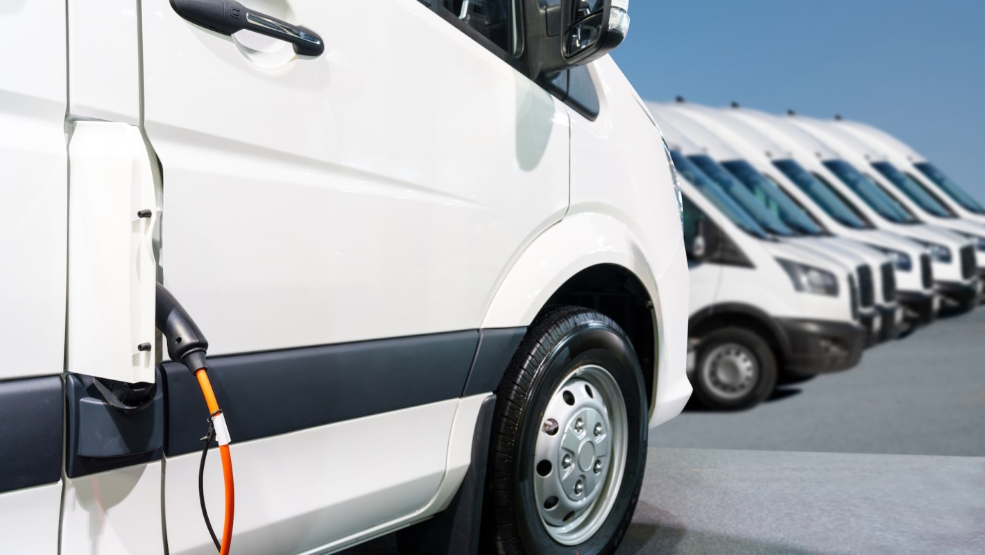 An electric van with a charging cable is parked among several white vans in a commercial setting.