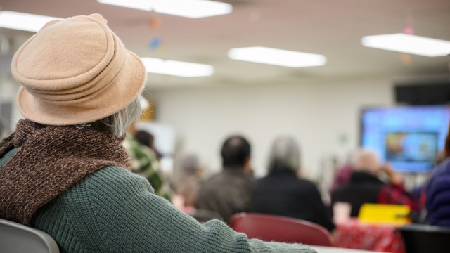 An elderly person in a pink hat sits facing a group watching a presentation in a communal space.