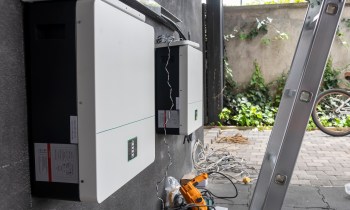 Two solar inverters mounted on a wall with nearby tools and cables, and greenery in the background.