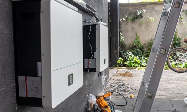 Two solar inverters mounted on a wall with nearby tools and cables, and greenery in the background.