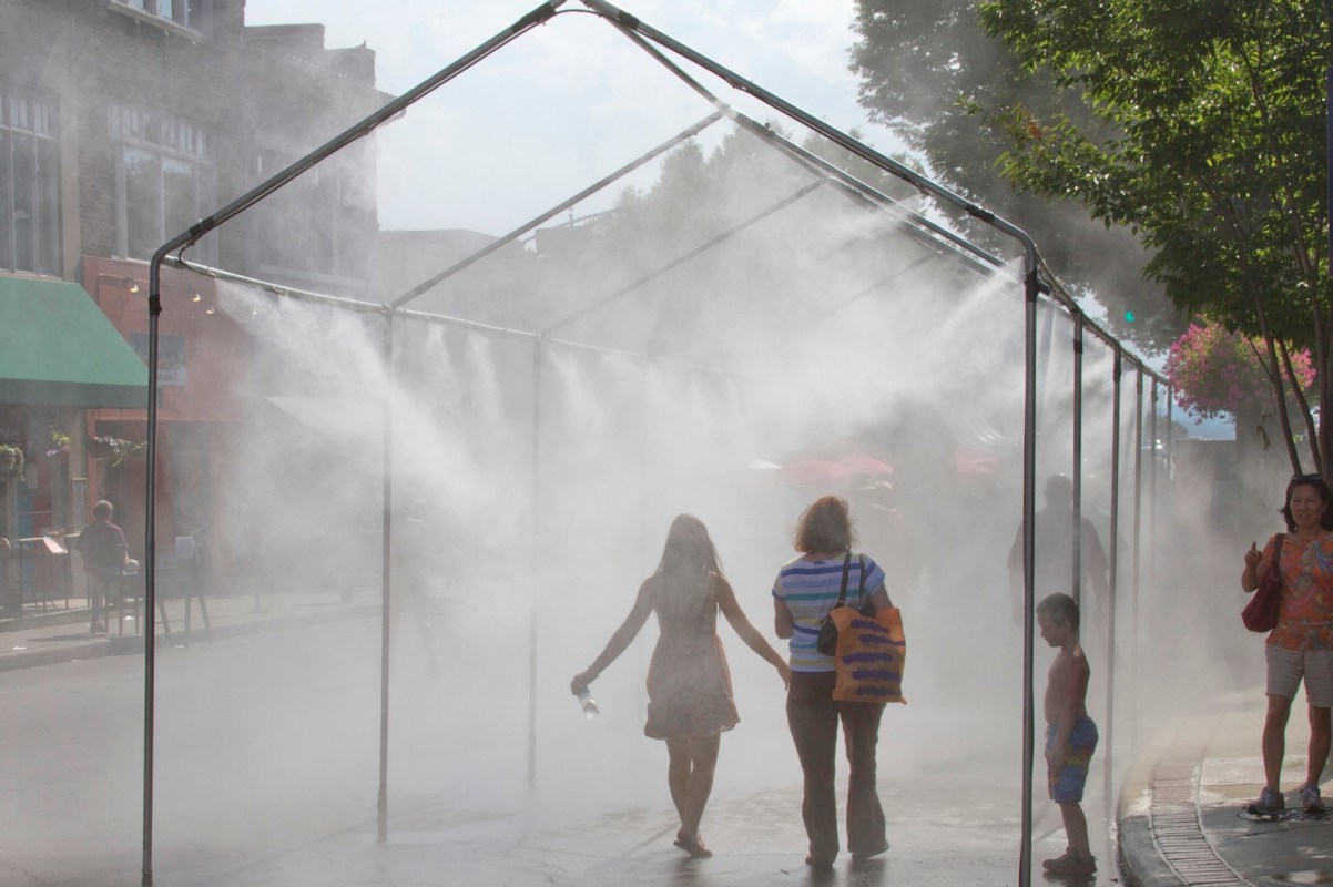 Two people walk through a misty water spray in a bustling street, with a child standing nearby.