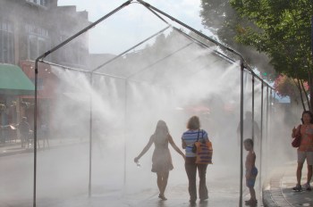 Two people walk through a misty water spray in a bustling street, with a child standing nearby.