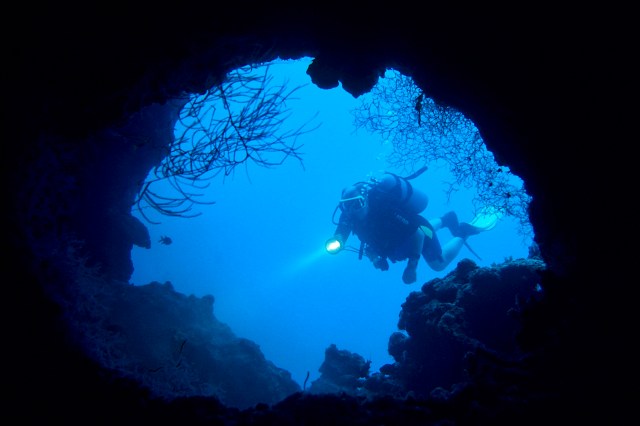 A scuba diver explores underwater near a rocky entrance illuminated by a flashlight.
