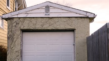 A small, weathered detached garage with a white door and textured yellow exterior, next to a wooden fence.