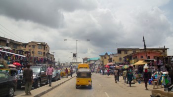 A busy street market scene with vendors, shoppers, and vehicles under a cloudy sky.