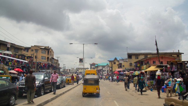 A busy street market scene with vendors, shoppers, and vehicles under a cloudy sky.