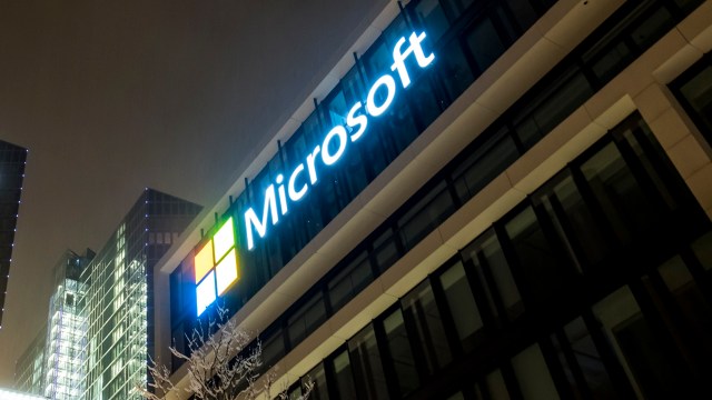 The Microsoft logo illuminated on a modern building during nighttime, surrounded by glass structures.