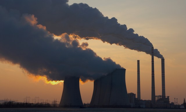 Smoke rises from cooling towers and chimneys from a coal plant against a sunset sky, symbolizing industrial activity.