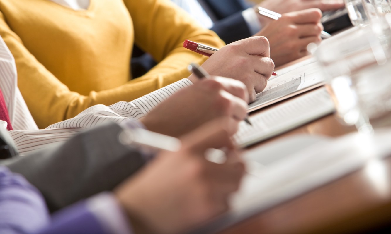 A close-up of right-handed people writing notes during a meeting, with a mix of casual and formal attire visible.