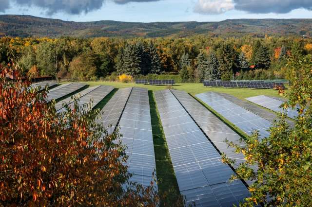 A solar panel field stretches across a grassy area, surrounded by colorful autumn foliage and rolling hills.
