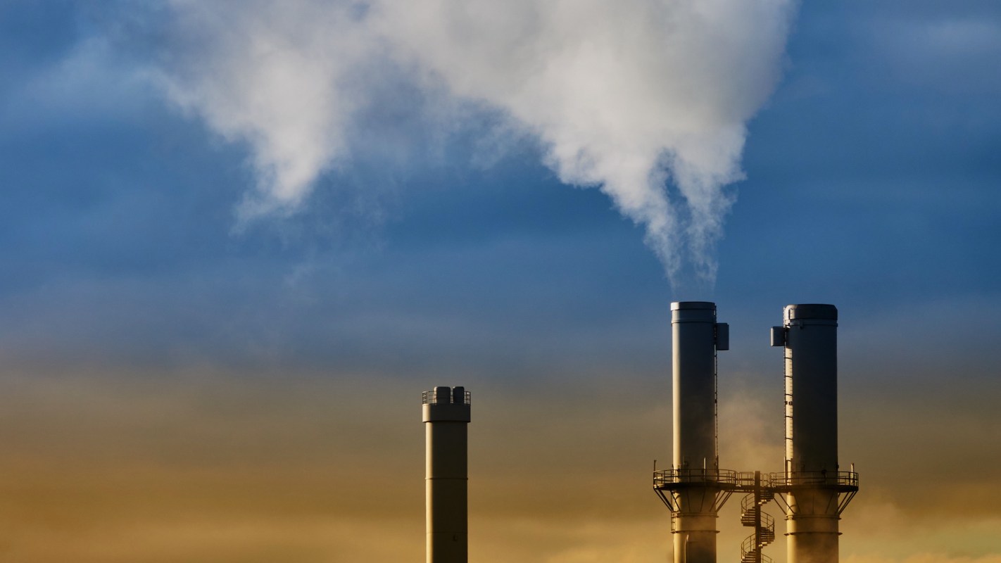 Two industrial smokestacks releasing white smoke against a blue sky, with a gradient of yellow and brown below.