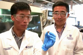 Two researchers in lab coats and safety glasses examine a small electronic component in a laboratory setting.