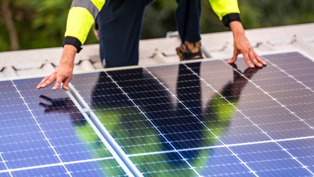 A worker in a reflective jacket installs solar panels on a rooftop under natural light.