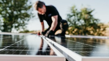 A worker installs solar panels on a rooftop under clear blue skies.