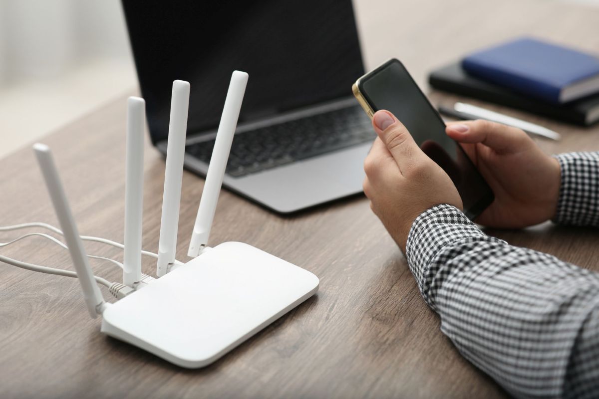 A person holds a smartphone next to a Wi-Fi router and a laptop.