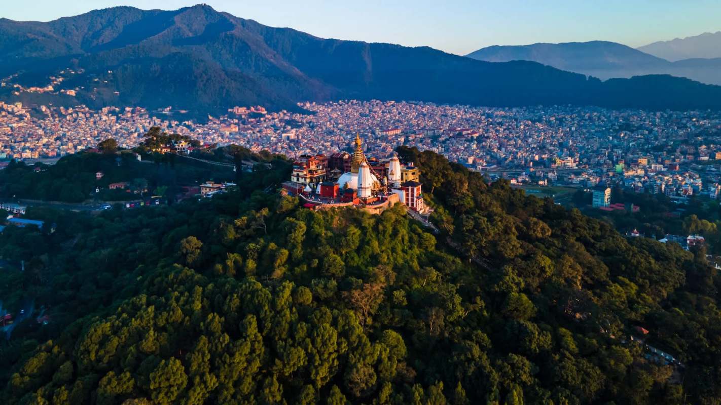 Aerial view of a temple complex atop a lush hill, overlooking a sprawling city and mountains in the background.