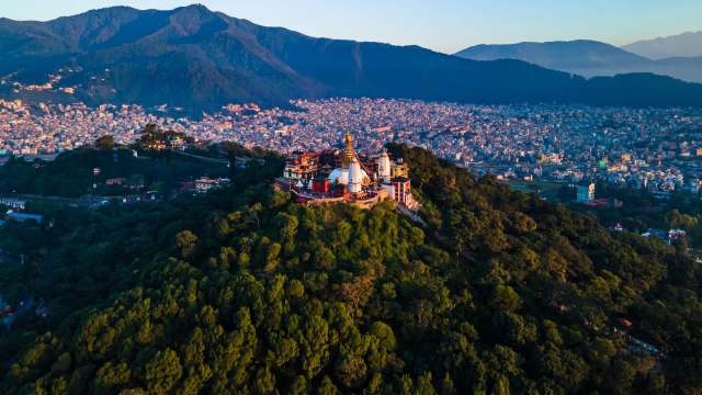 Aerial view of a temple complex atop a lush hill, overlooking a sprawling city and mountains in the background.