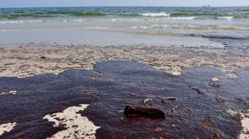Oil spills on a sandy beach, with waves in the background and a small piece of debris on the shore.