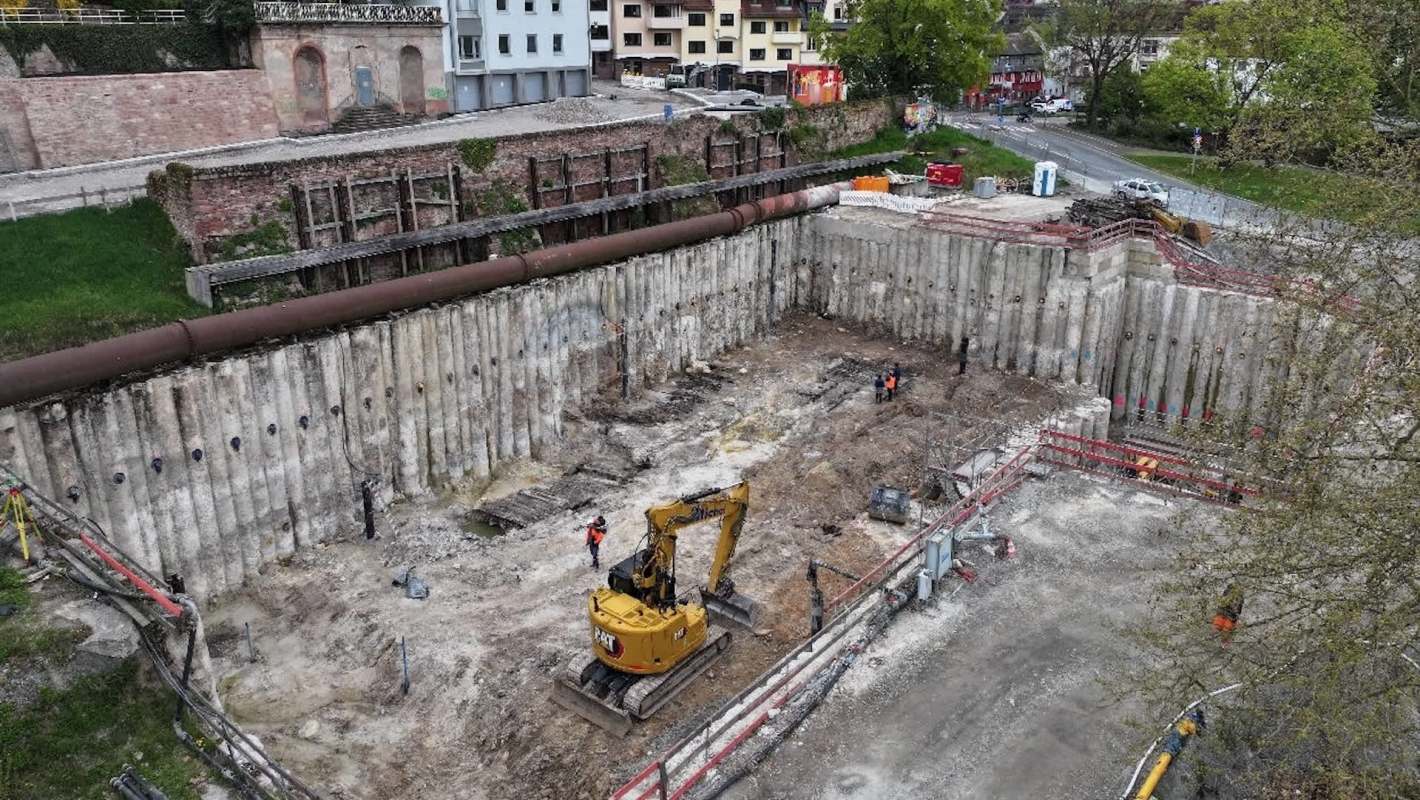 An excavation site with construction workers and machinery, surrounded by concrete walls and a grassy area.
