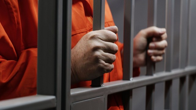 A close-up of hands gripping metal bars, wearing an orange prison jumpsuit.