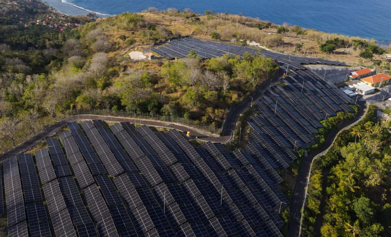 A solar panel installation on a hillside near the ocean, surrounded by greenery and houses.