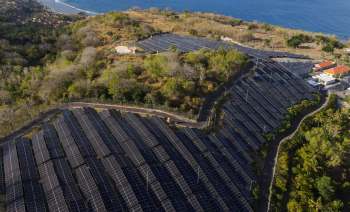 A solar panel installation on a hillside near the ocean, surrounded by greenery and houses.