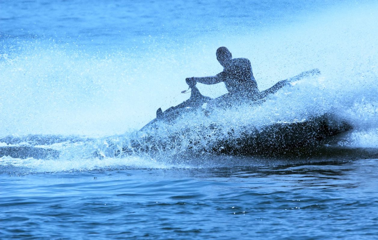 A silhouette of a person riding a jet ski through splashing waves on a calm body of water.