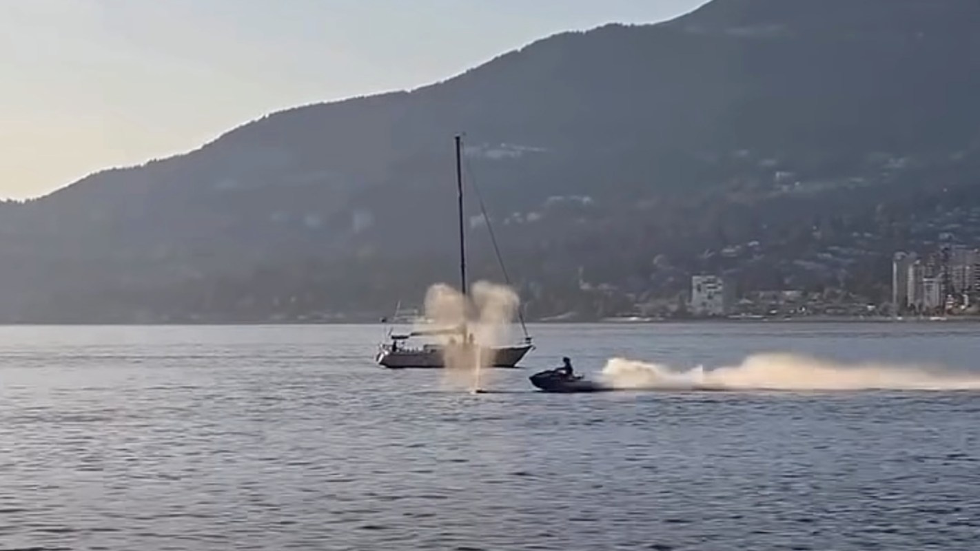 A jet ski speeds across the water near a sailboat, with mountains and buildings in the background.