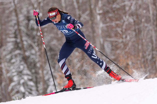 A competitive skier in a blue racing suit gracefully strides through the snowy landscape, creating a spray of snow.