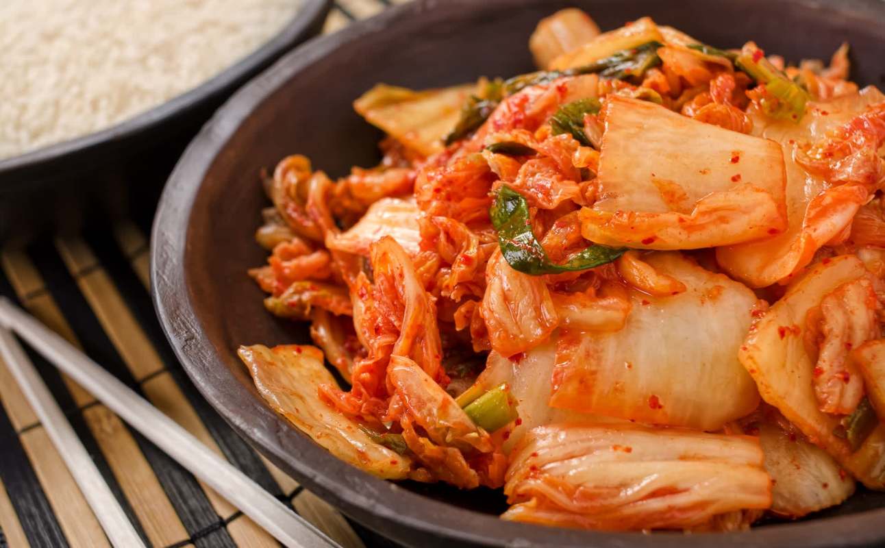 A bowl of kimchi with vibrant red chili and green onions, served alongside a bowl of white rice.