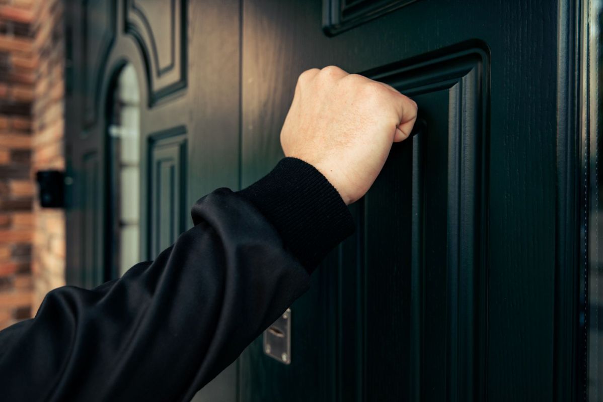 A hand in a black sweater is knocking on a dark green door.