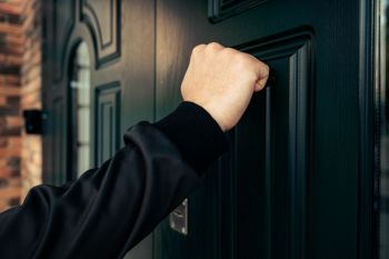A hand in a black sweater is knocking on a dark green door.
