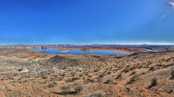 A panoramic view of a desert landscape with a calm blue lake and distant mesas under a clear blue sky.