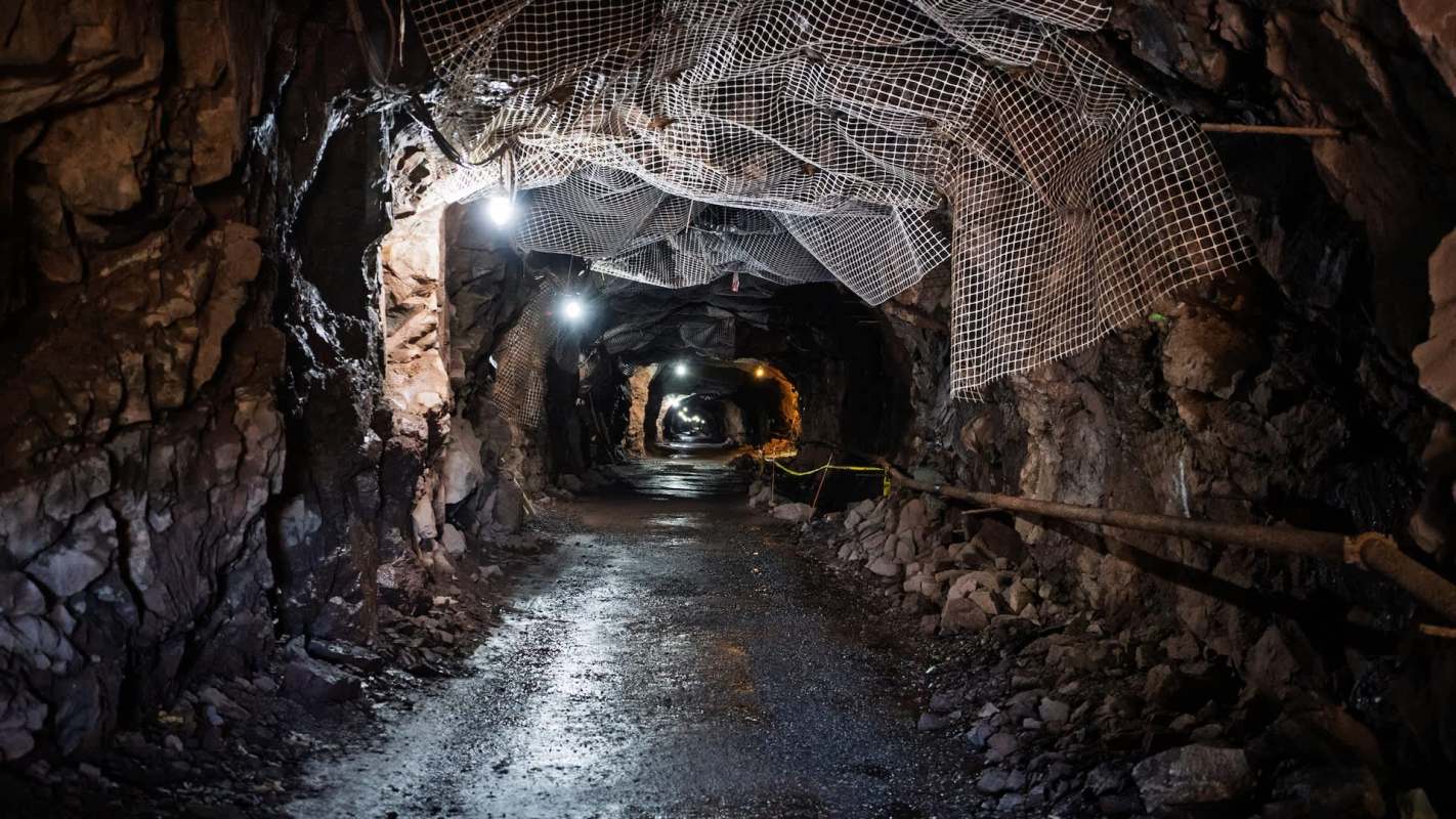 A dimly lit underground mine tunnel with rocky walls and support netting above.