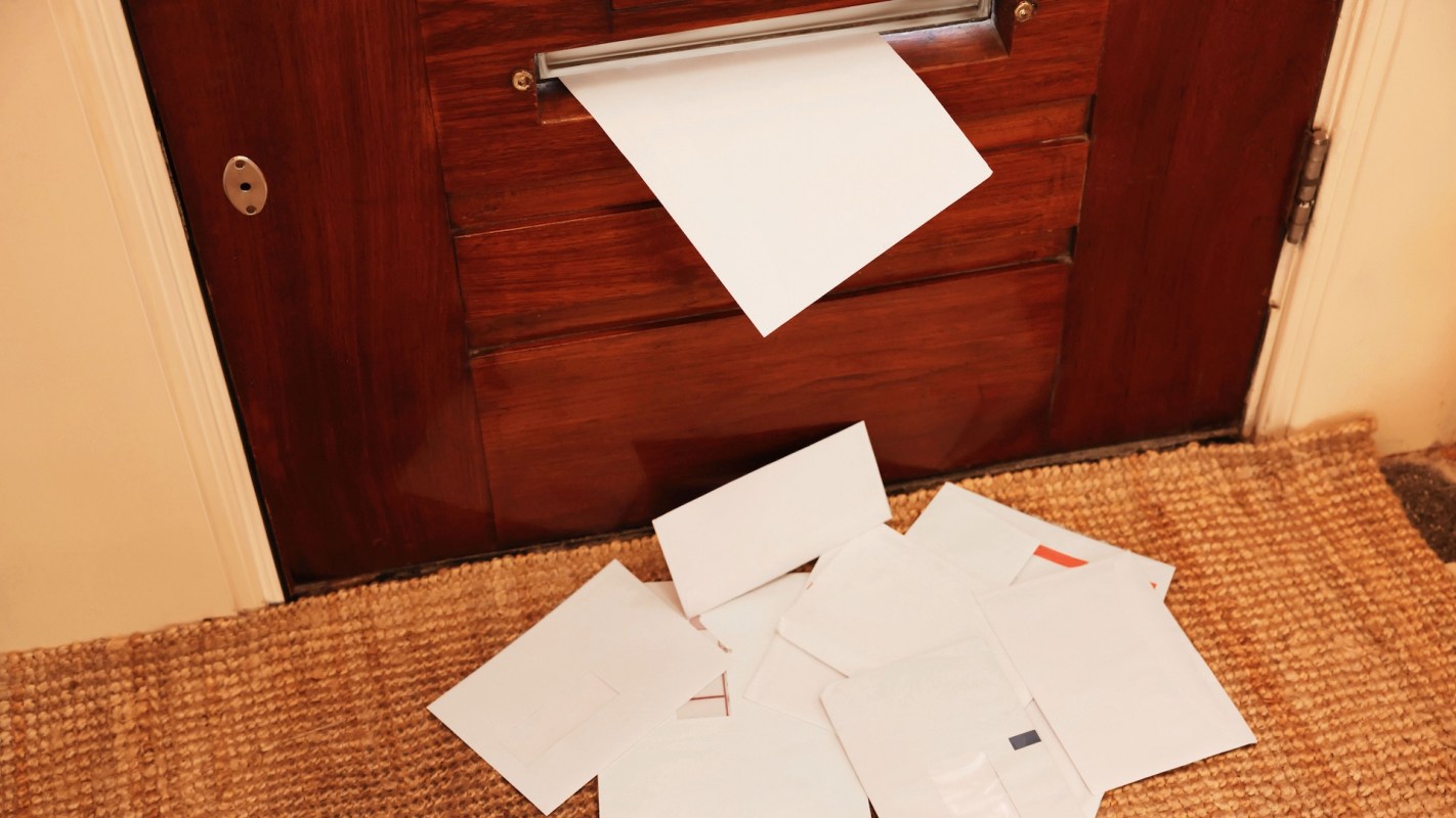 A pile of letters scattered on a doormat with one letter partly sticking out of a mail slot in a wooden door.