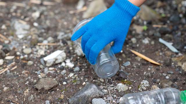 A hand wearing a blue glove picks up a clear plastic cup from a dirt and gravel ground.