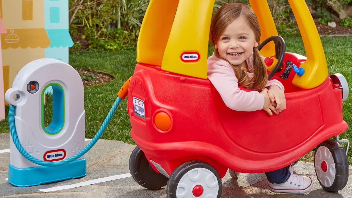 A smiling child sits in a red toy car next to a play electric pump in a grassy outdoor setting.